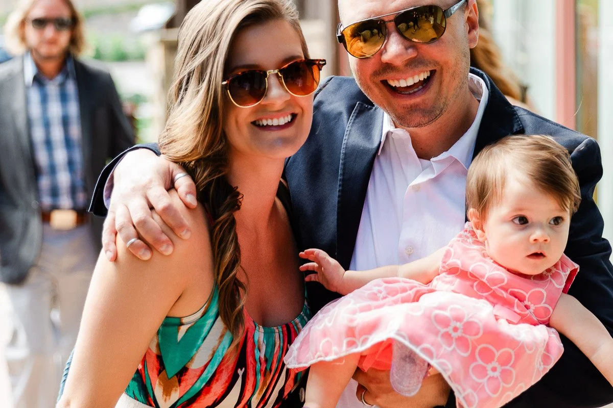 Smiling couple in sunglasses pose with a baby in a pink dress. The man holds the baby, and a casually-dressed man walks behind them in a sunny setting.