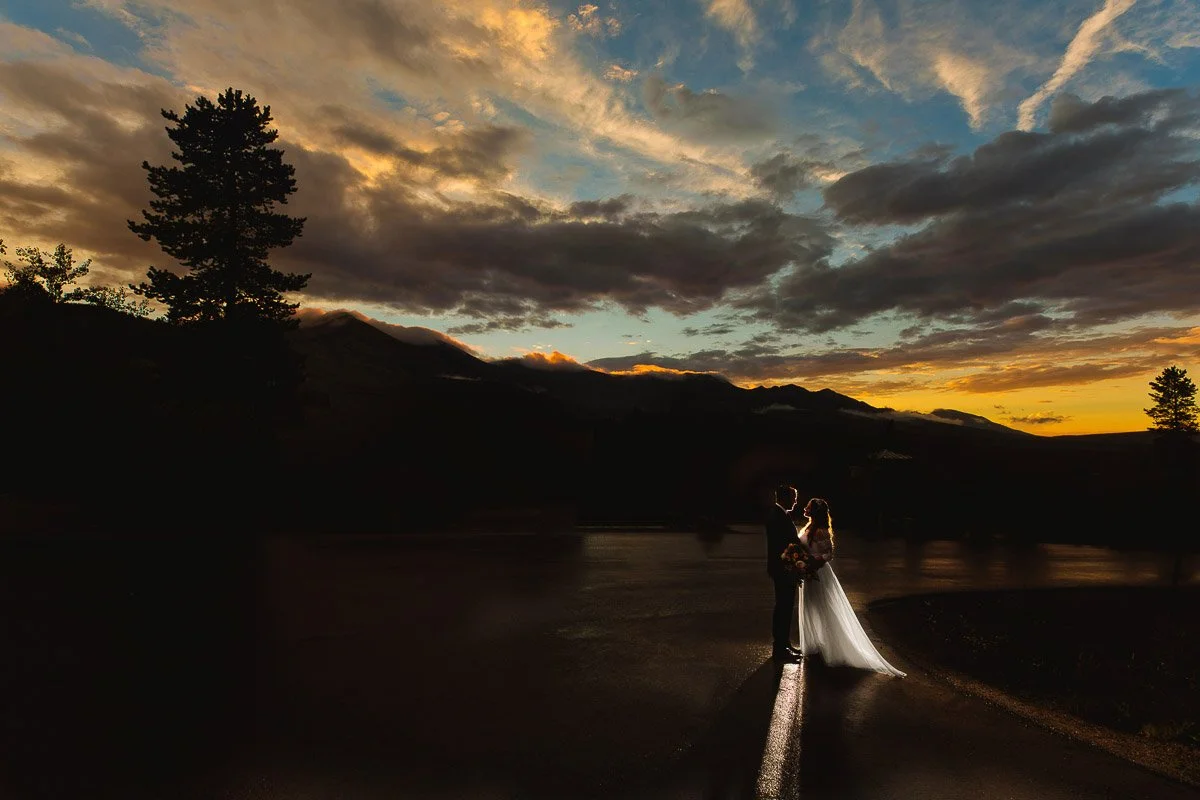 A couple stands embracing on a road during sunset, silhouetted against a dramatic sky with vivid orange and blue hues, creating a romantic atmosphere captured by Breckenridge wedding photographer tomKphoto