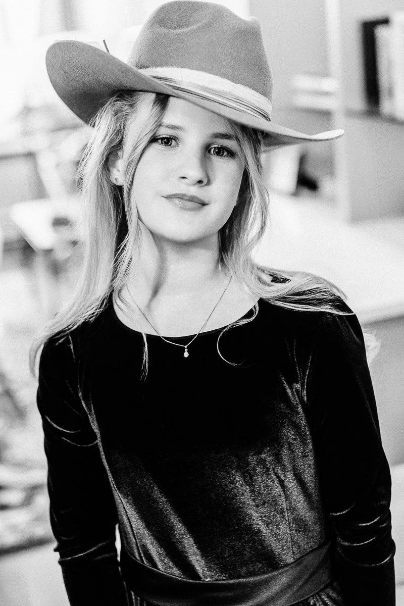 Young girl in a cowboy hat and velvet dress poses confidently indoors. Black-and-white image conveys a timeless, nostalgic feel.