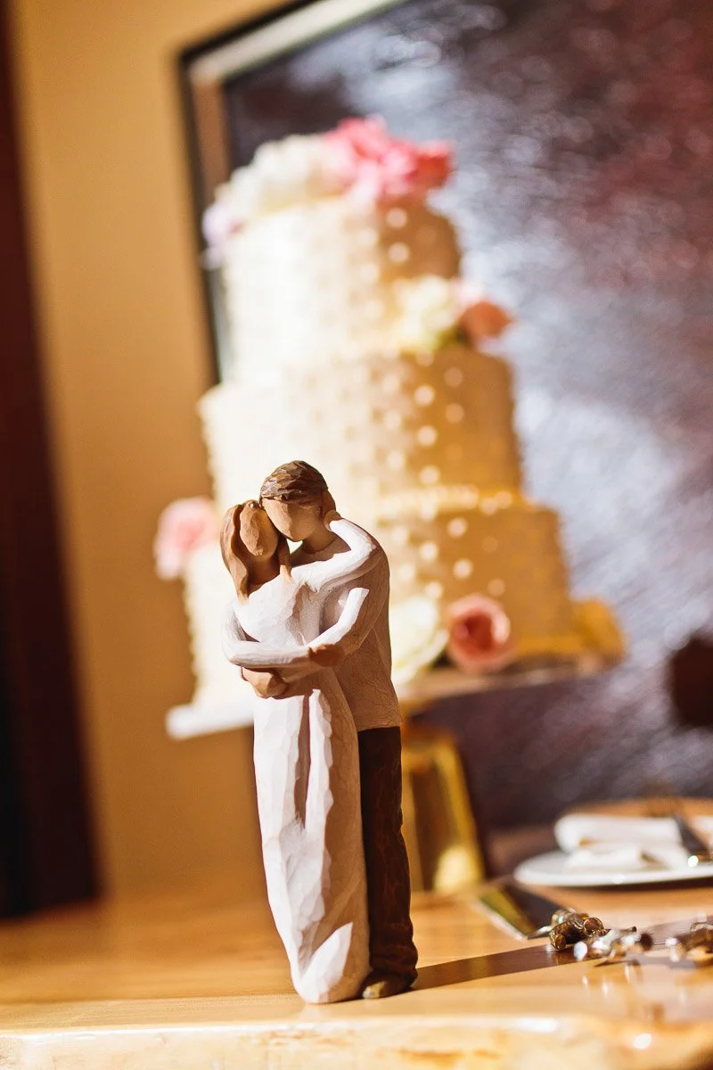 Rustic figurine of an embracing couple in the foreground, with a softly lit, three-tiered wedding cake adorned with roses blurred in the background. Warm, romantic atmosphere.