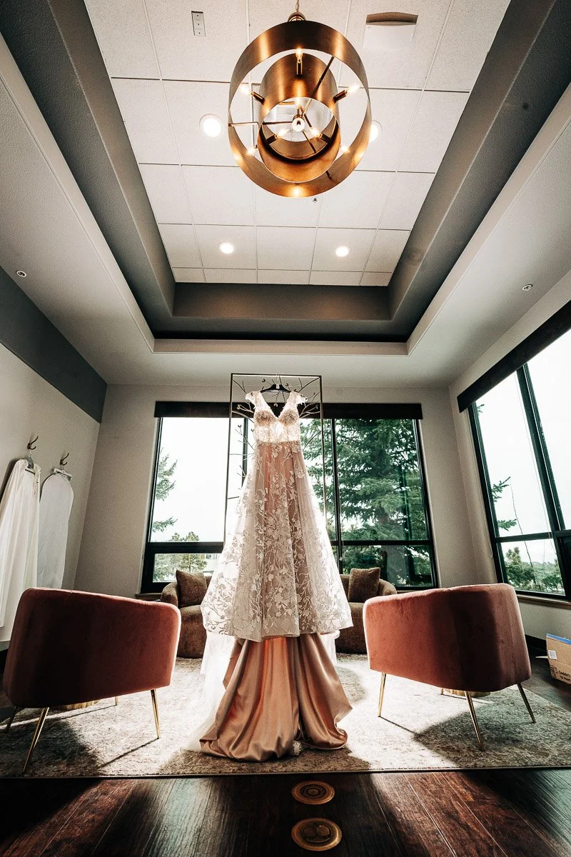 A lace wedding dress on a mannequin in an elegant room with large windows, pink chairs, and a gold chandelier, creating a luxurious and serene atmosphere.