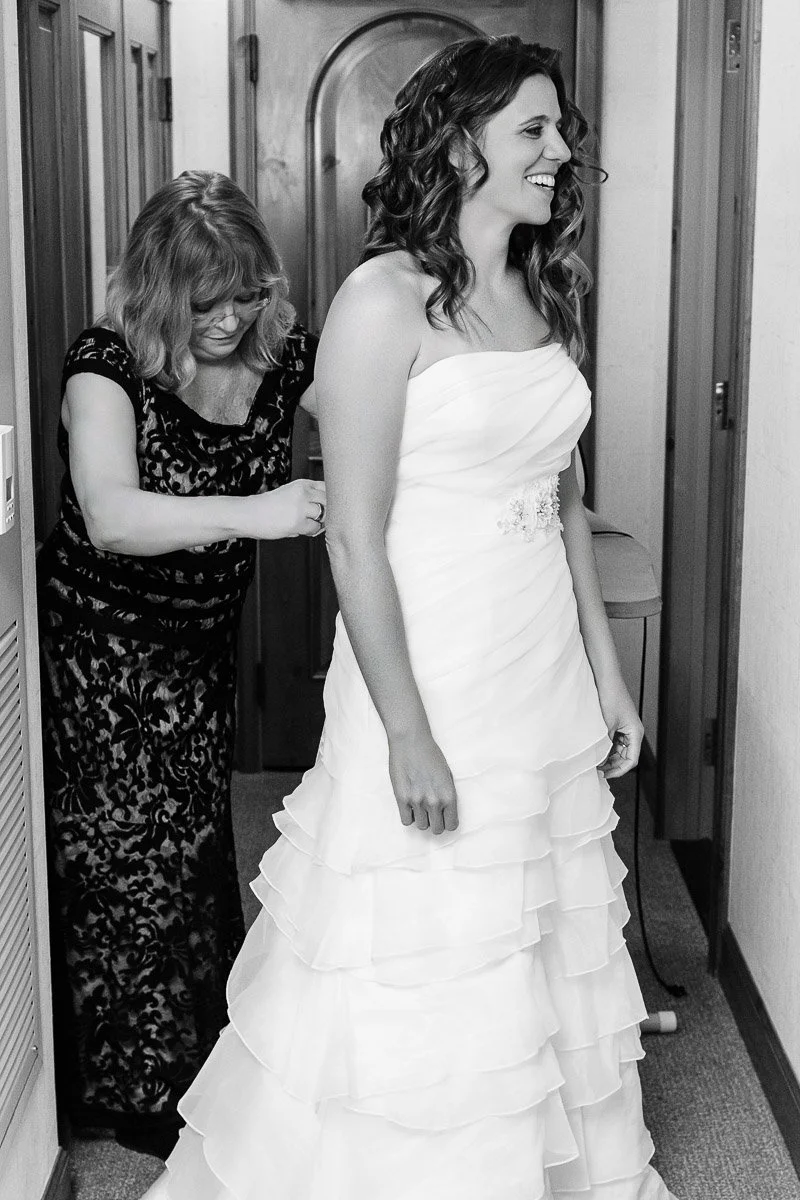 A woman in a white wedding dress smiles as another woman helps fasten her gown in a hallway. The scene is joyful and intimate, highlighting a bonding moment.