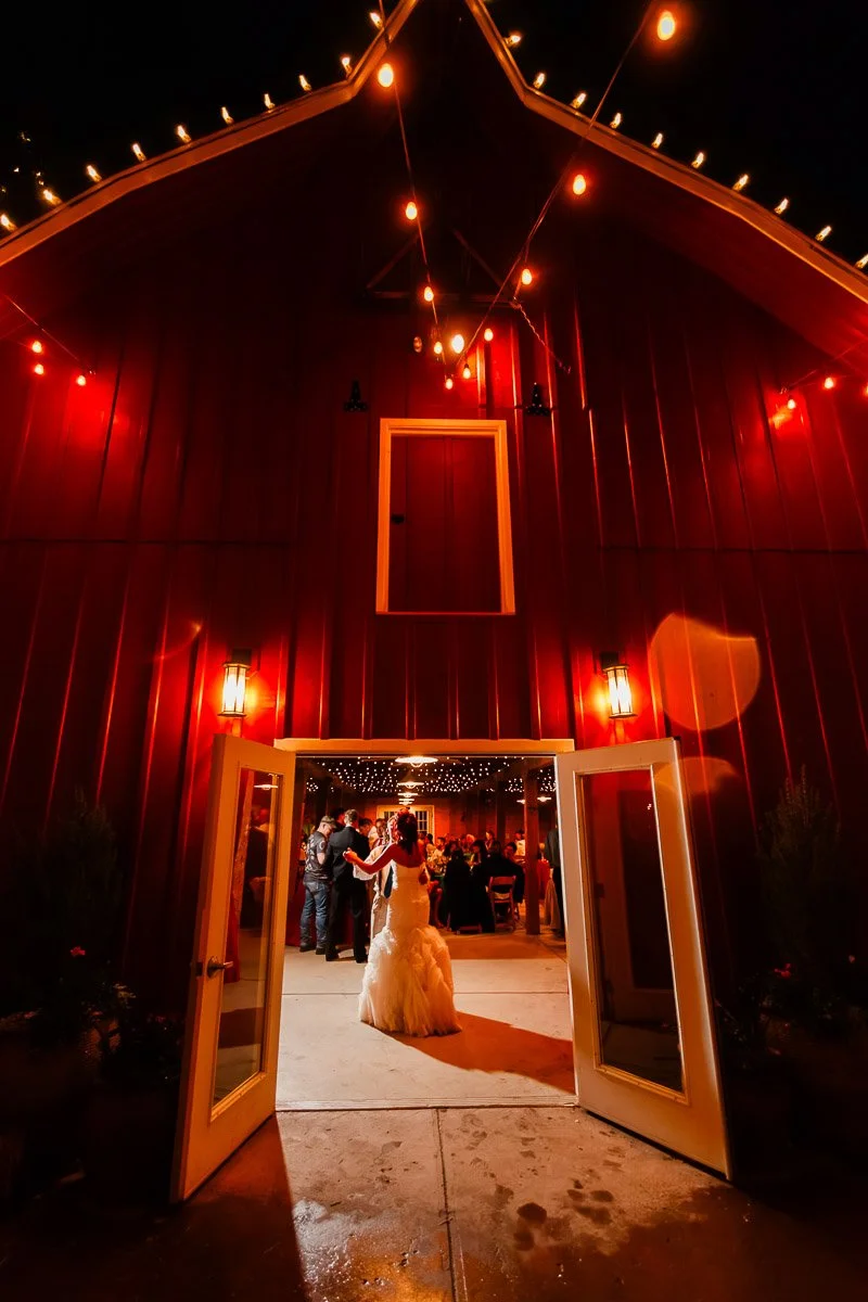 A bride in a white gown stands at the entrance of a warmly lit red barn adorned with string lights. Inside, guests gather, creating a festive atmosphere.