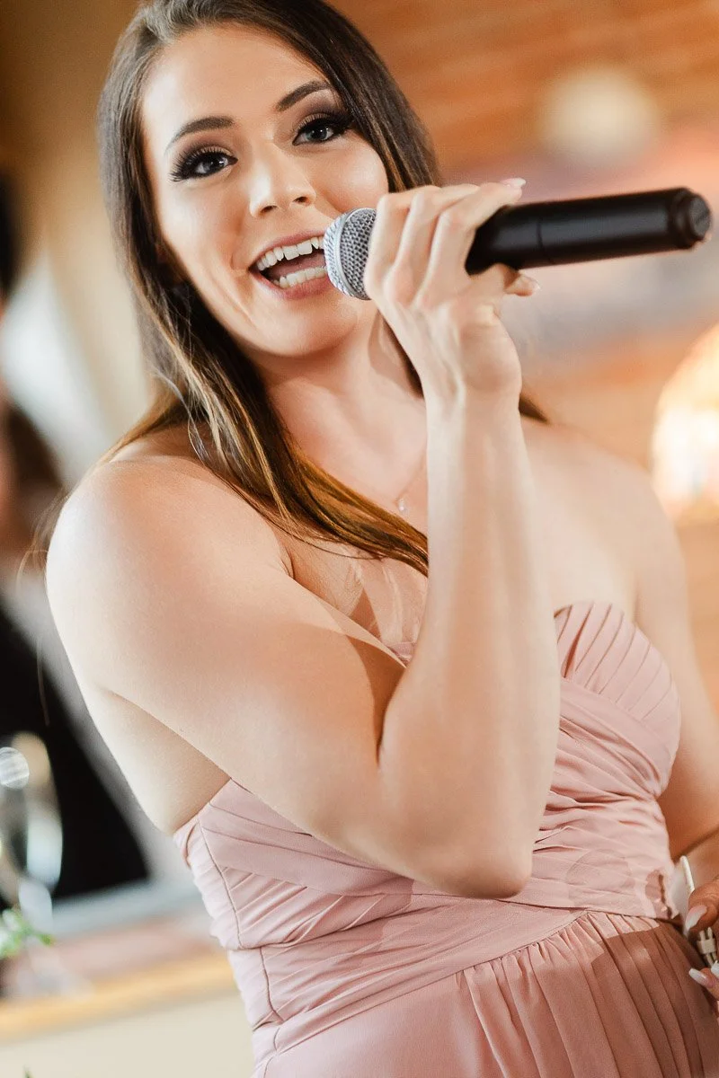 A woman in a strapless pink dress holds a microphone, smiling as she speaks. The background is softly blurred, suggesting a lively, warm atmosphere.