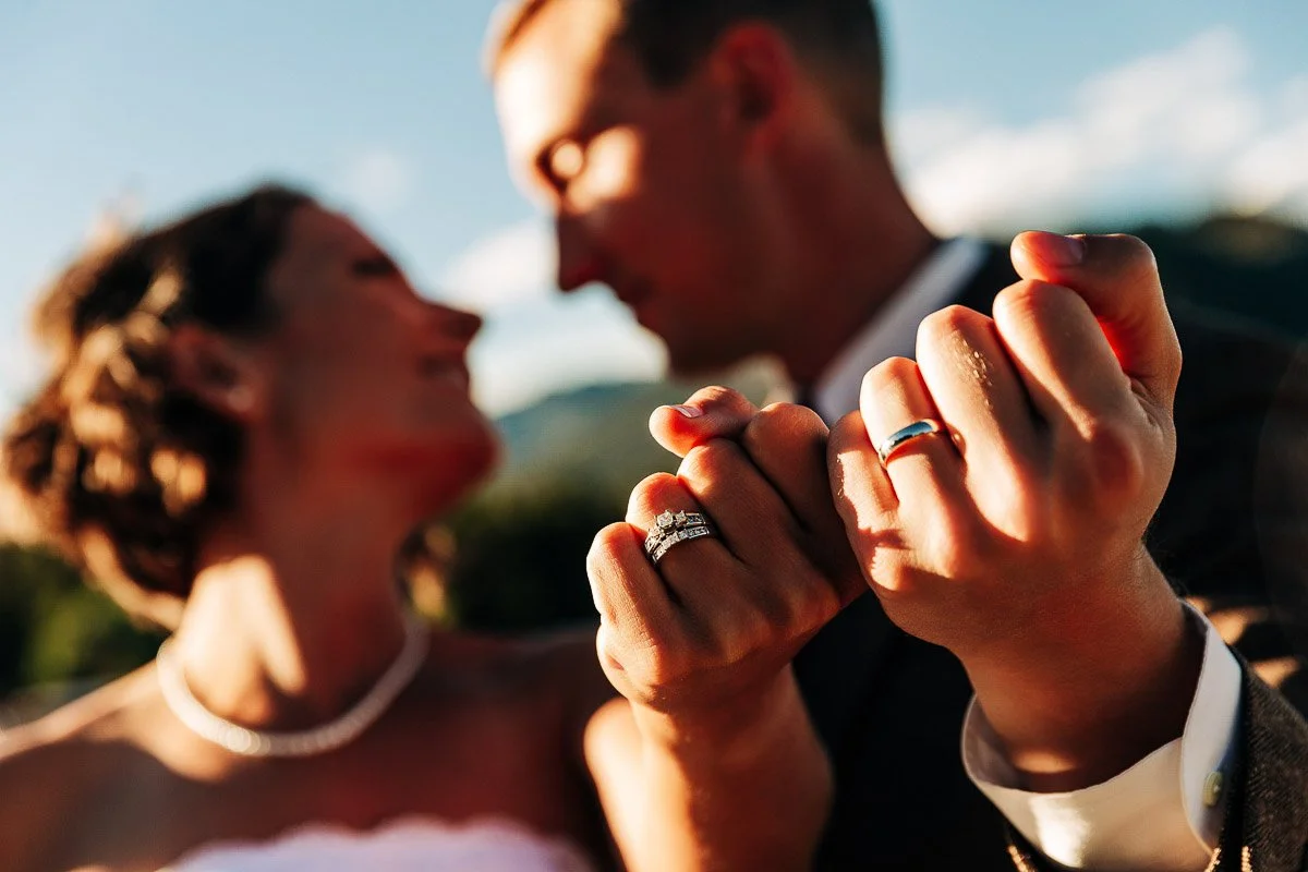 A couple in wedding attire focus on their interlocked hands, displaying wedding rings. Soft light and a blurred nature backdrop create a romantic ambiance.
