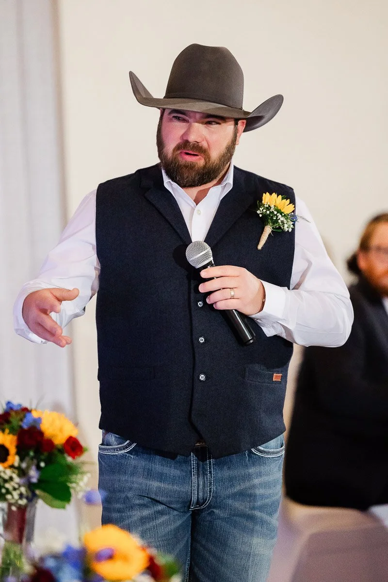 A man in a cowboy hat and vest holds a microphone mid-speech, gesturing with one hand. He wears a flower boutonniere, with a colorful flower arrangement nearby.