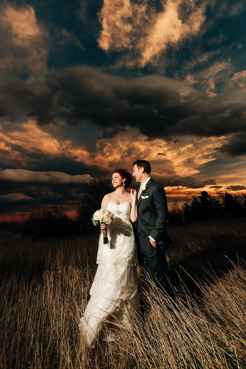 Bride in a lace gown and groom in a suit stand in a field at sunset, holding a bouquet. The dramatic sky casts a romantic, warm glow during a Greenbriar Inn wedding in Boulder, Colorado.