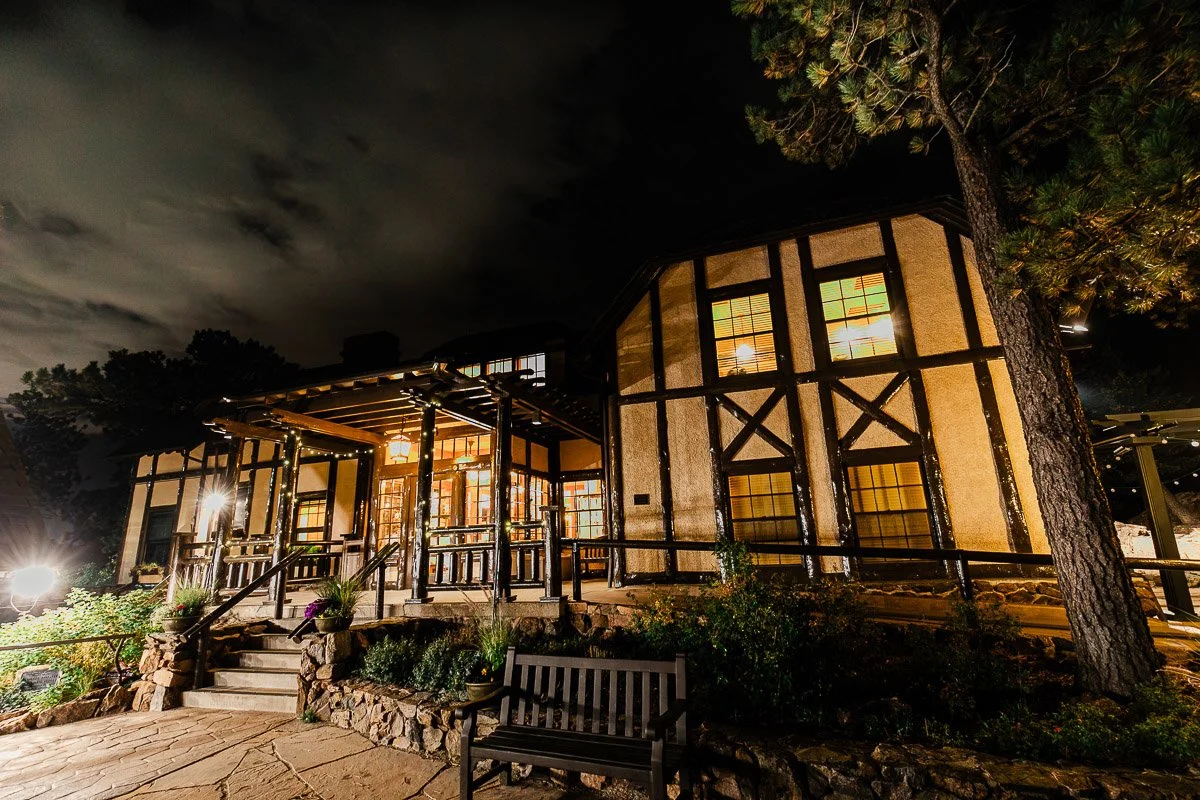 Nighttime view of an illuminated, rustic house with timber framing. Warm lights glow from windows. A bench and stone path are in the foreground.