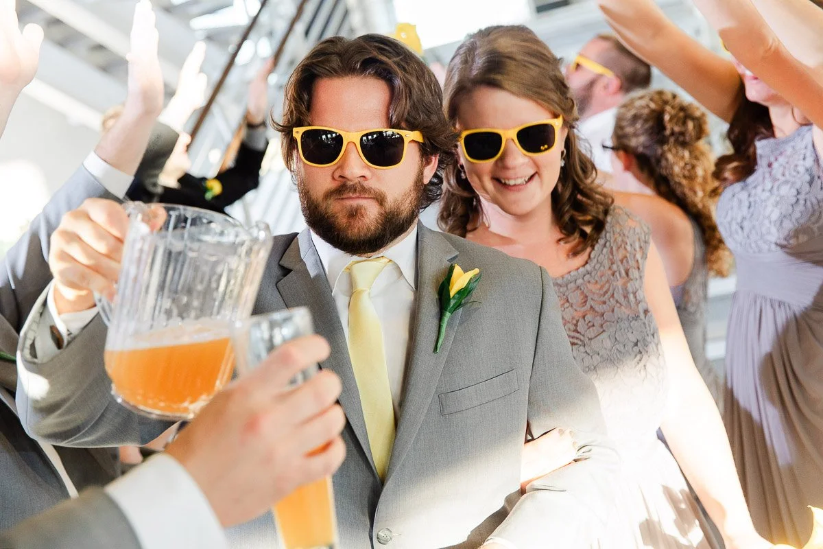 A man in a gray suit and a woman in a lace dress, both wearing yellow sunglasses, smile as they walk through a celebratory tunnel of friends holding drinks.