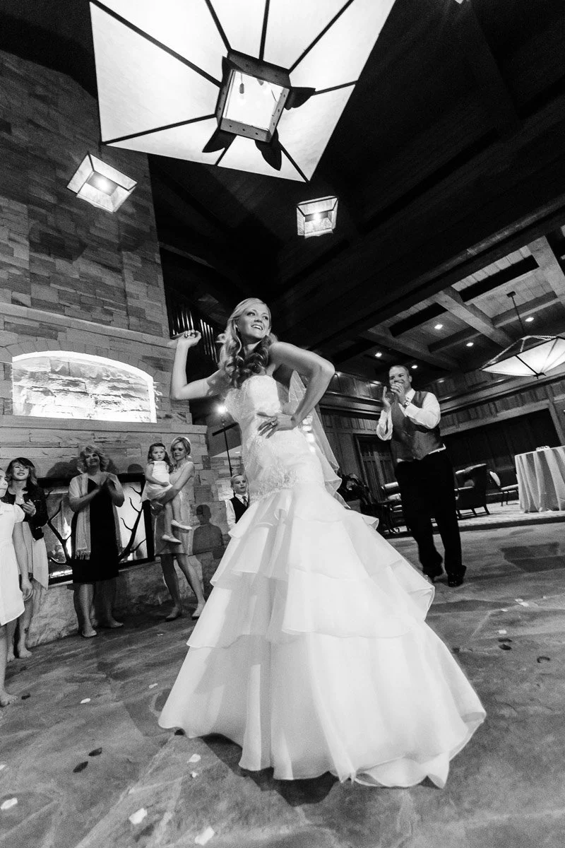 A bride joyfully dances in a flowing gown in a rustic venue. Onlookers, clapping and smiling, gather near a stone fireplace, creating a festive atmosphere.