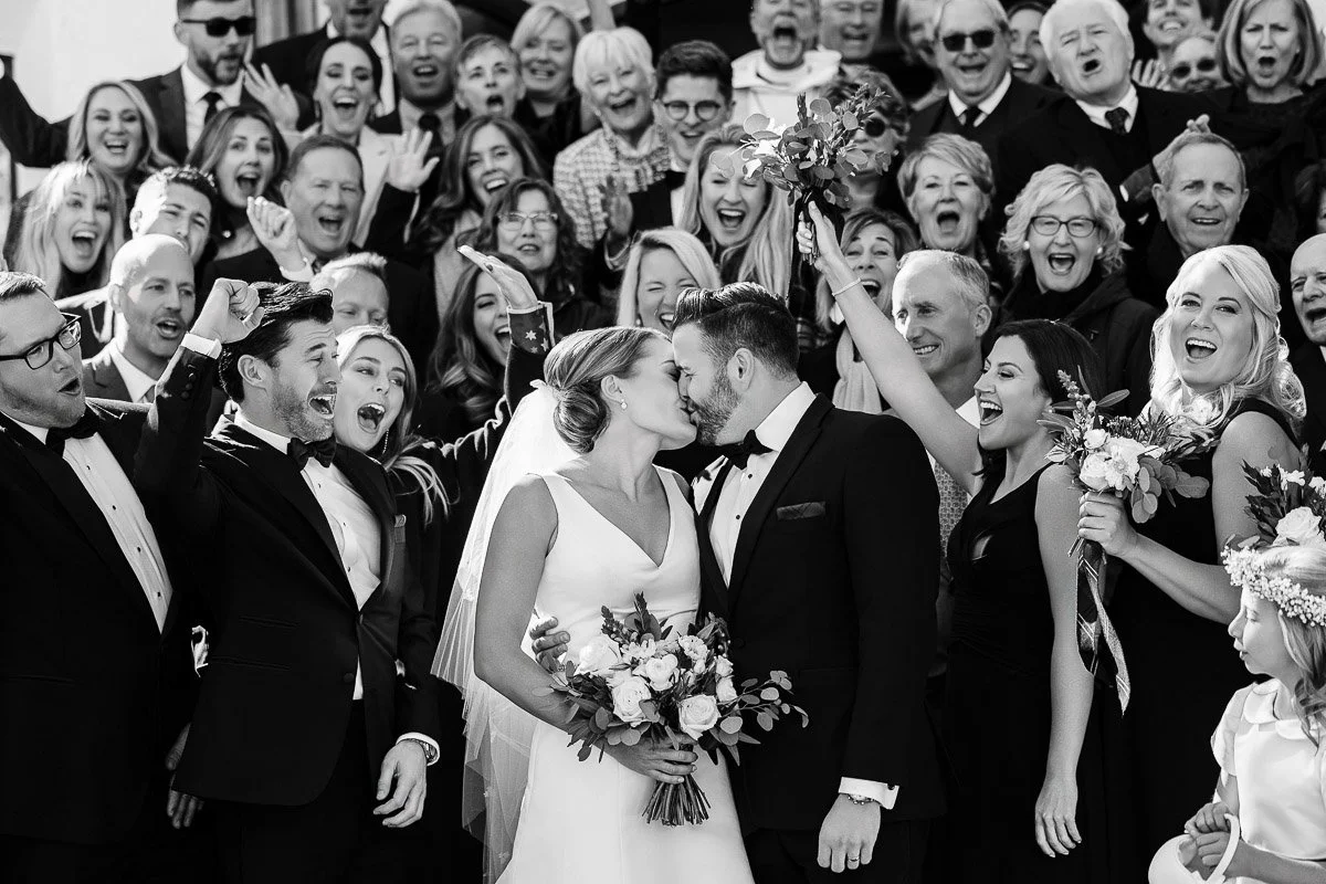 Bride and groom kiss joyfully amid cheering guests at a wedding. The bride holds a bouquet; bridesmaids and groomsmen celebrate. Festive and emotional.