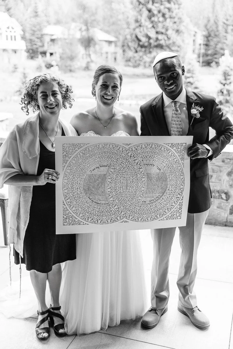 Black and white photo of three people at a wedding. The bride and groom hold a decorative marriage certificate. All are smiling, standing outdoors.