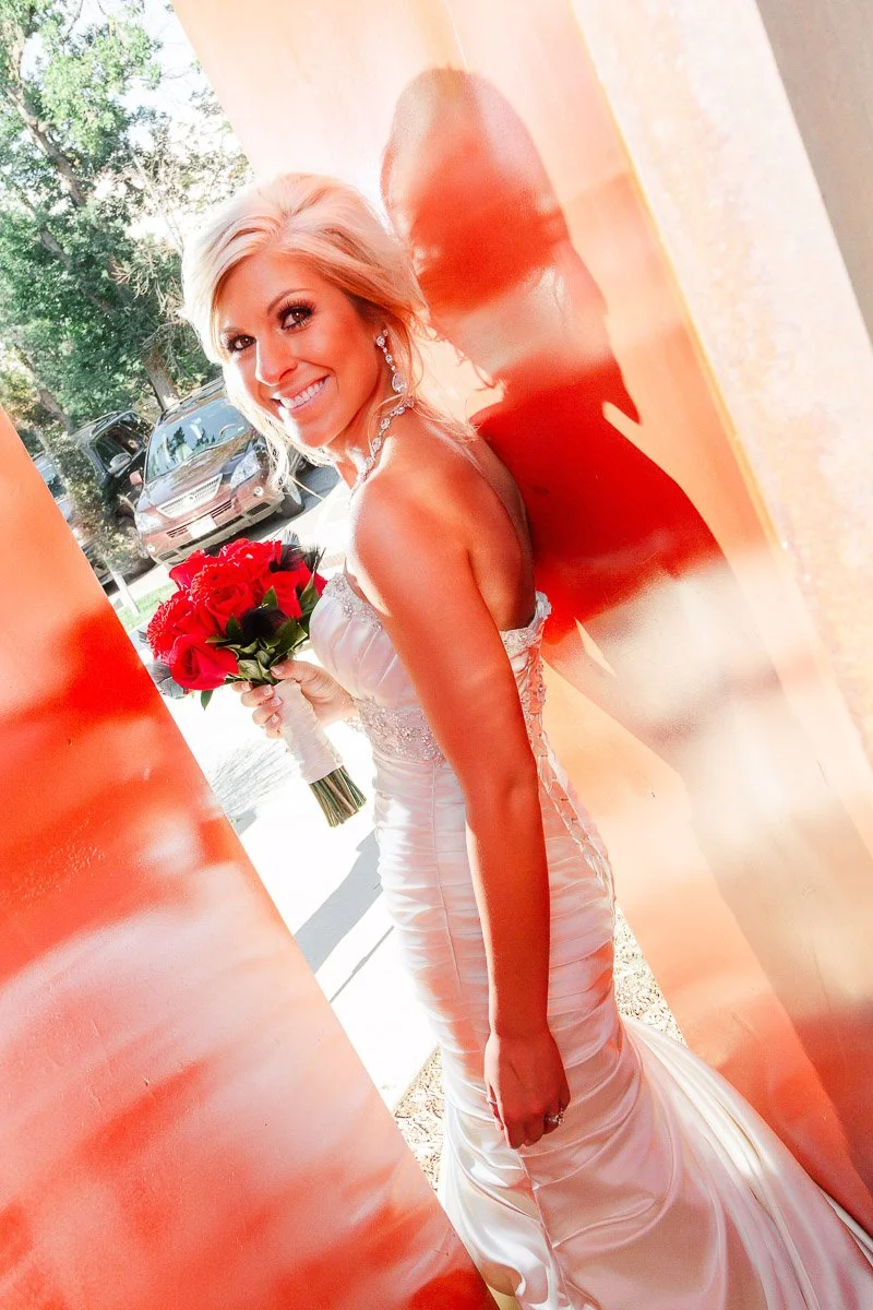 Bride in a white gown smiles brightly, holding a bouquet of red roses. She stands against an orange wall, casting a soft shadow. Sunlit and joyful scene.