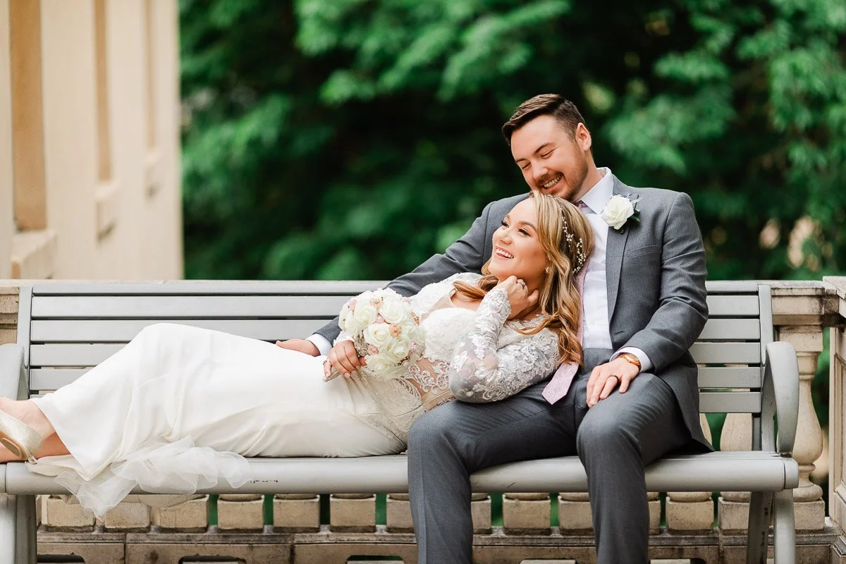 A bride in a white gown and a groom in a gray suit sit joyfully on a bench. The bride holds a bouquet, with greenery in the background, exuding romance during a GRANT HUMPHREYS MANSION in Denver, Colorado.