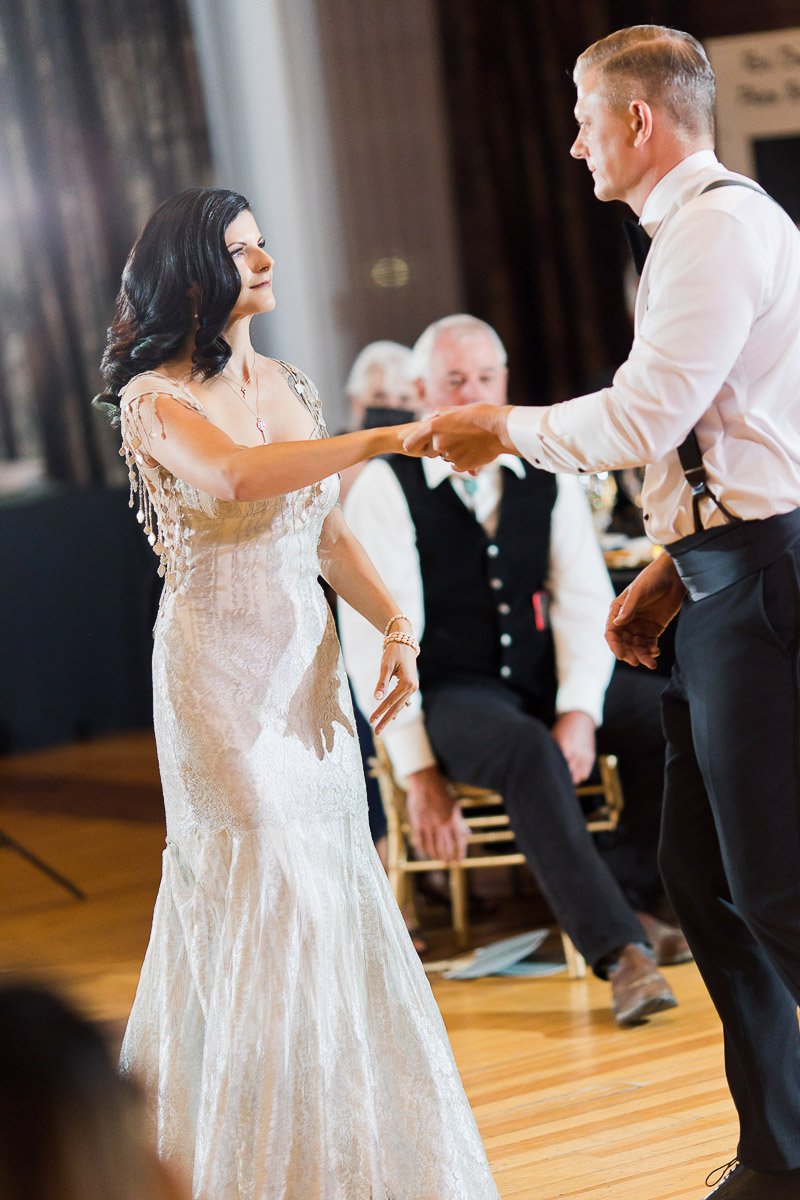A woman in an elegant white gown dances with a man in a white shirt and suspenders. The scene is warm and celebratory, with people seated in the background.