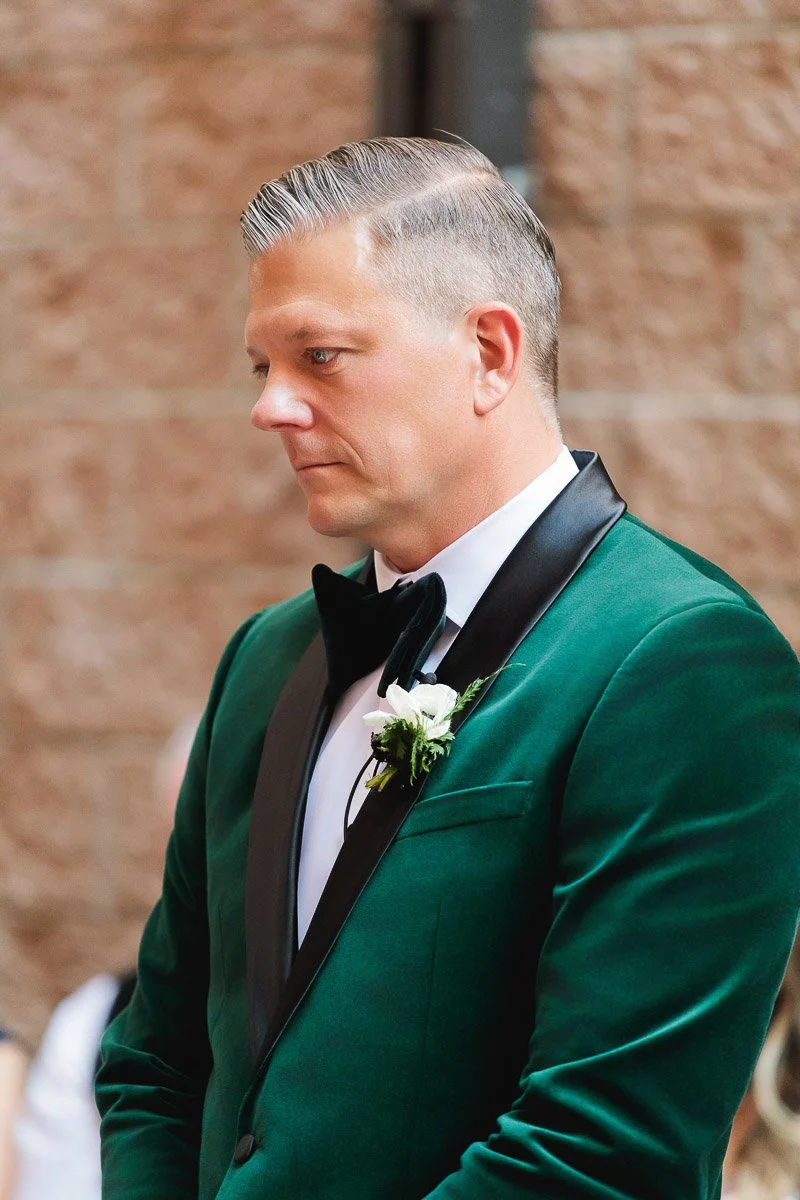Man in profile wearing a green velvet tuxedo with black lapels and a white boutonniere, looking thoughtful against a blurred brick wall background.