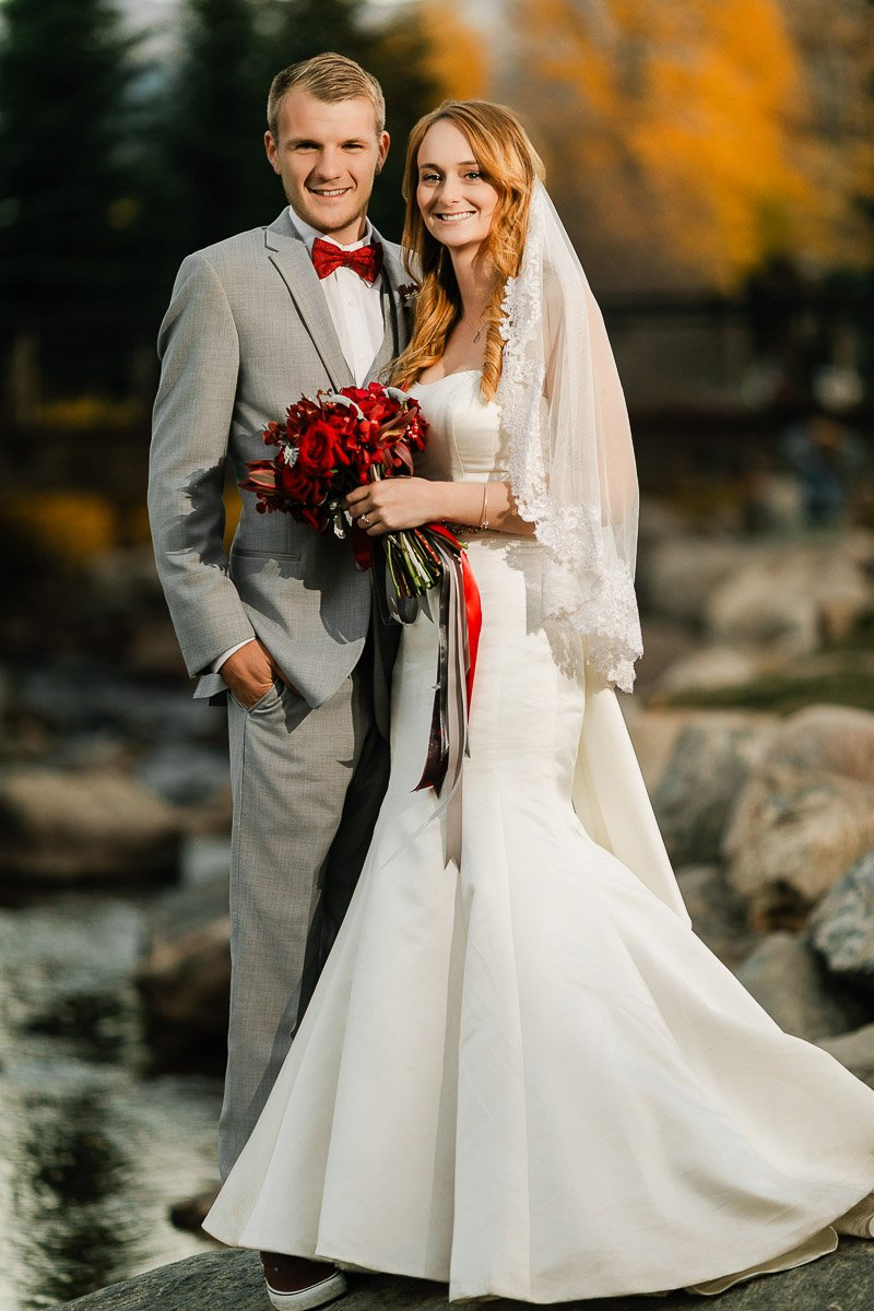 Bride and groom smiling in outdoor setting. Bride in white gown holds red bouquet; groom in gray suit and red bow tie. Autumn leaves in background.