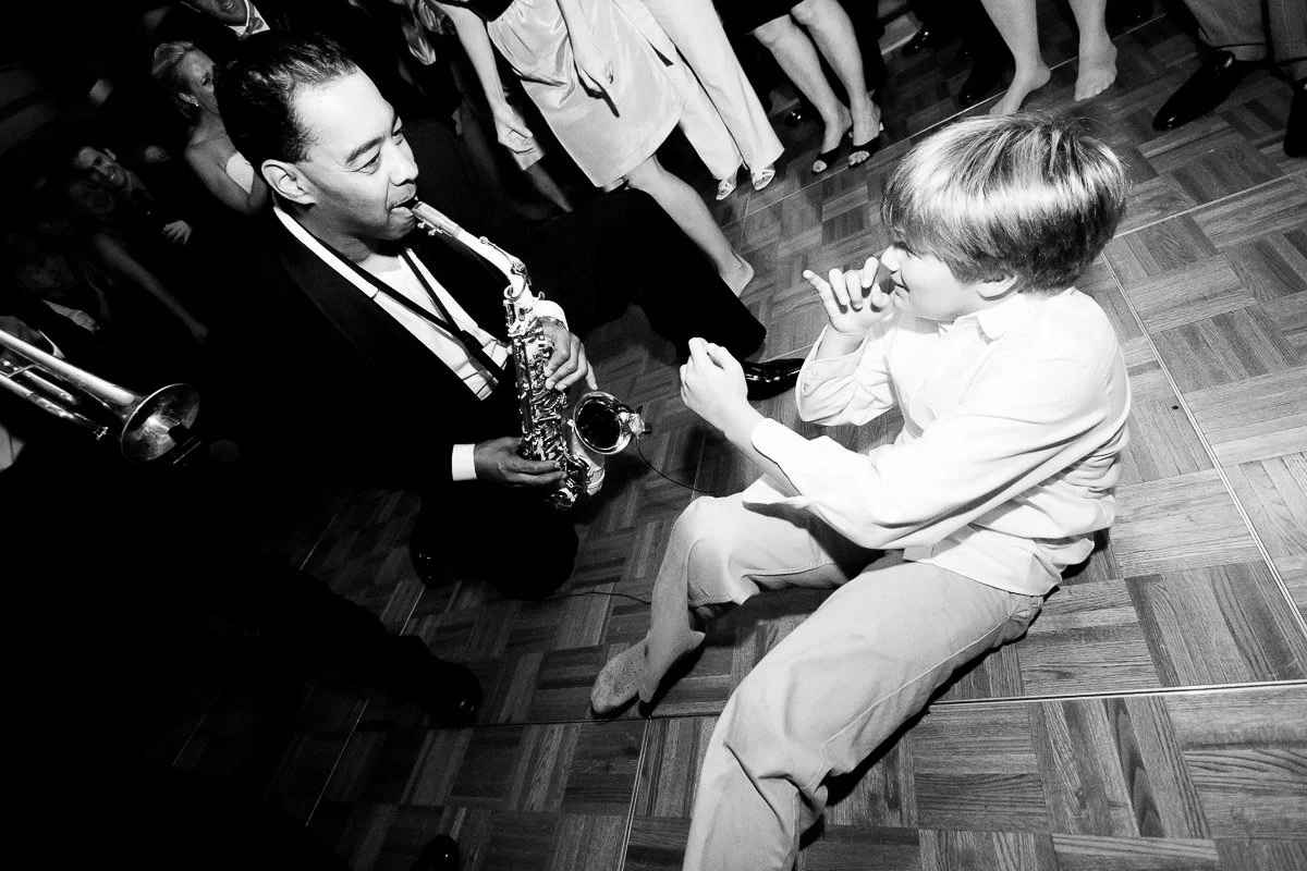 A musician playing saxophone kneels on a dance floor, engaging with a young boy who sits and mimics playing. Guests stand around, creating a joyful atmosphere.