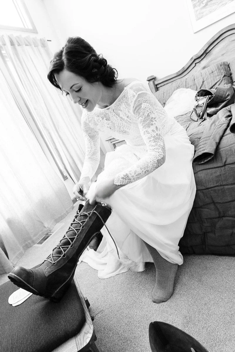 A bride in a lace wedding dress sits on a bed, joyfully putting on tall lace-up boots. The room is softly lit with a warm, intimate atmosphere.