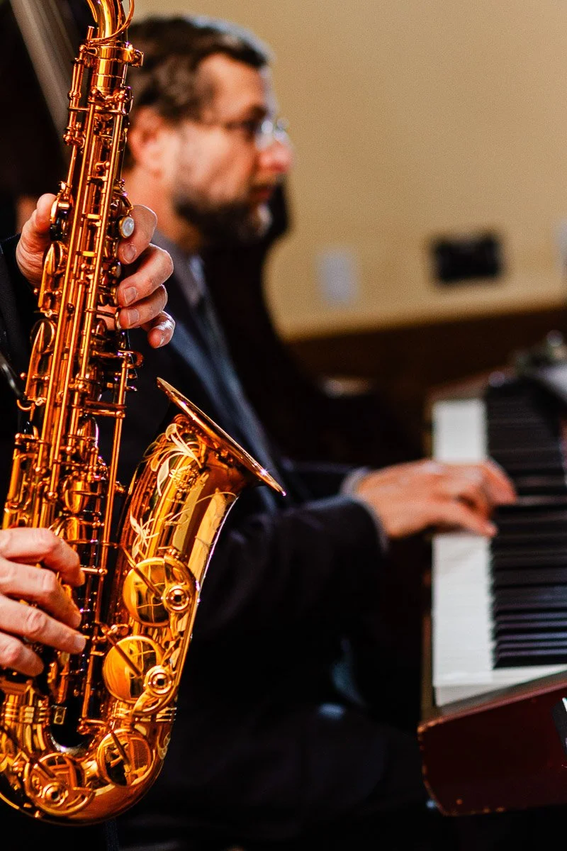 Close-up of a musician's hands playing a golden saxophone, with a blurred piano player in the background. The scene conveys a warm, jazzy ambiance.