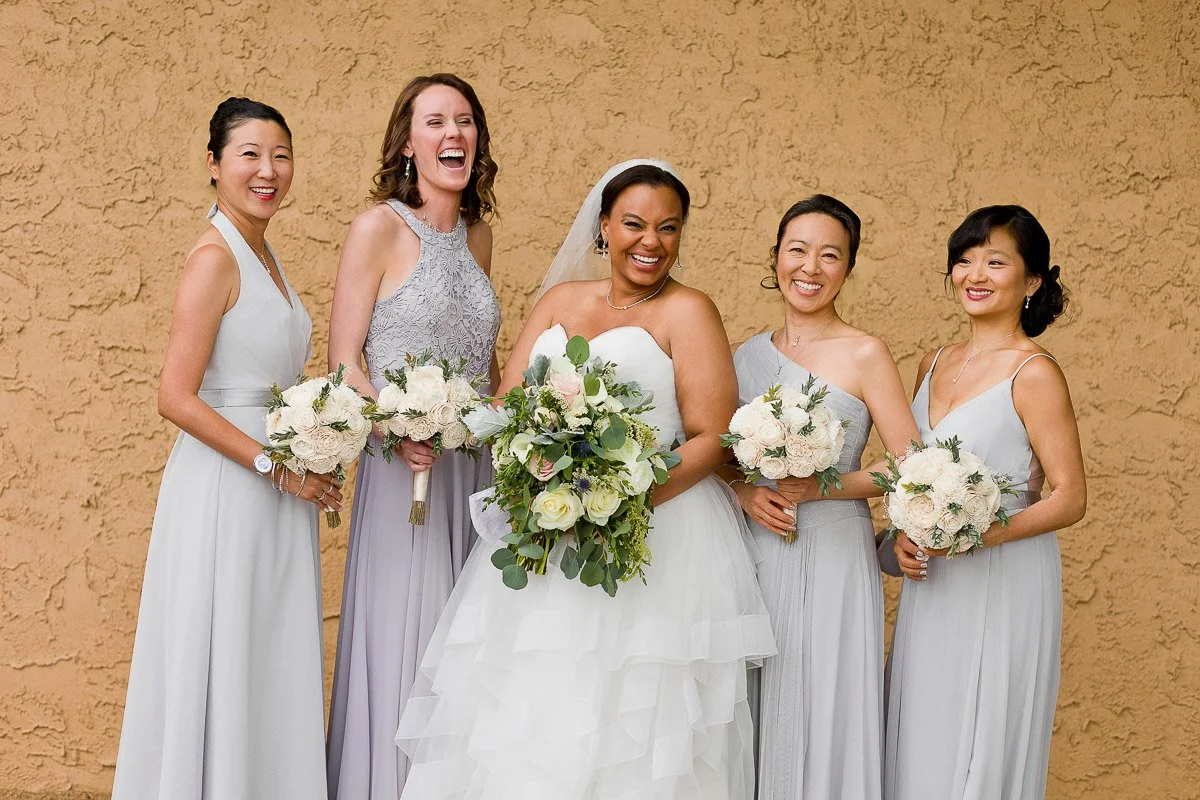 A joyful bride in a white gown stands with four bridesmaids in gray dresses, all smiling and holding bouquets against a textured beige wall.