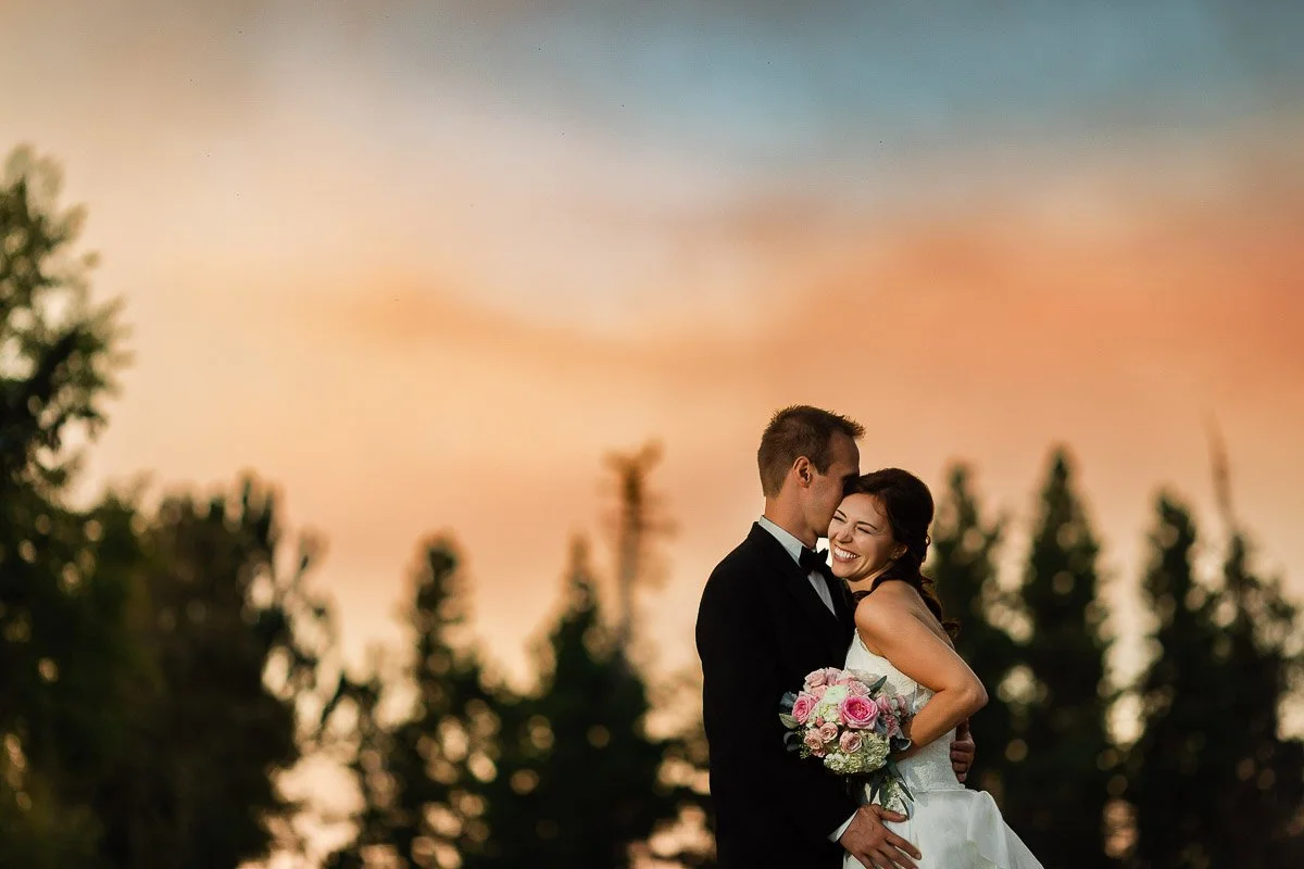 A couple, dressed in wedding attire, embrace joyfully under a vivid sunset sky with trees silhouetted in the background. The bride holds a bouquet of pink flowers.