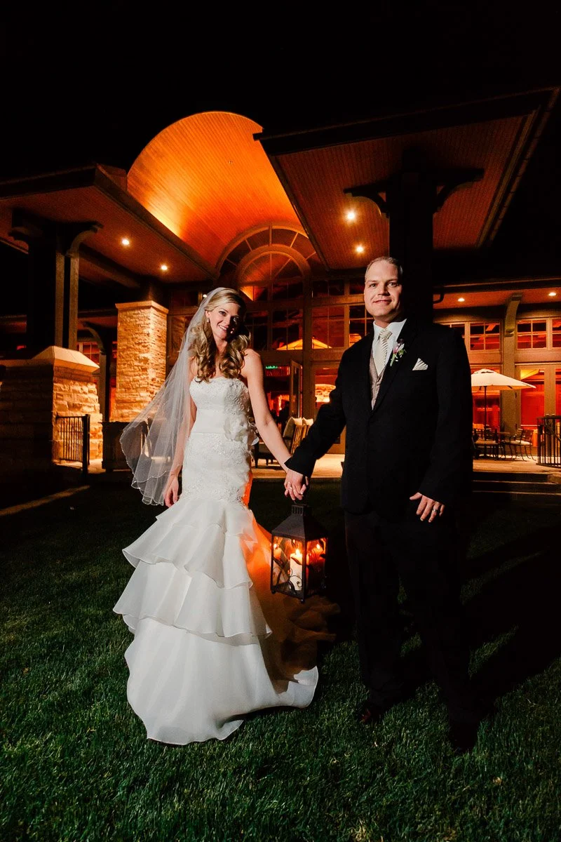 Bride in white gown and veil stands beside groom in black suit, holding hands at night. They hold a lantern, with a warmly lit building behind them.