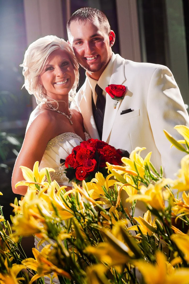 Smiling bride and groom pose joyfully indoors, surrounded by vibrant yellow lilies. Bride holds red rose bouquet; groom wears white suit with red rose boutonniere.