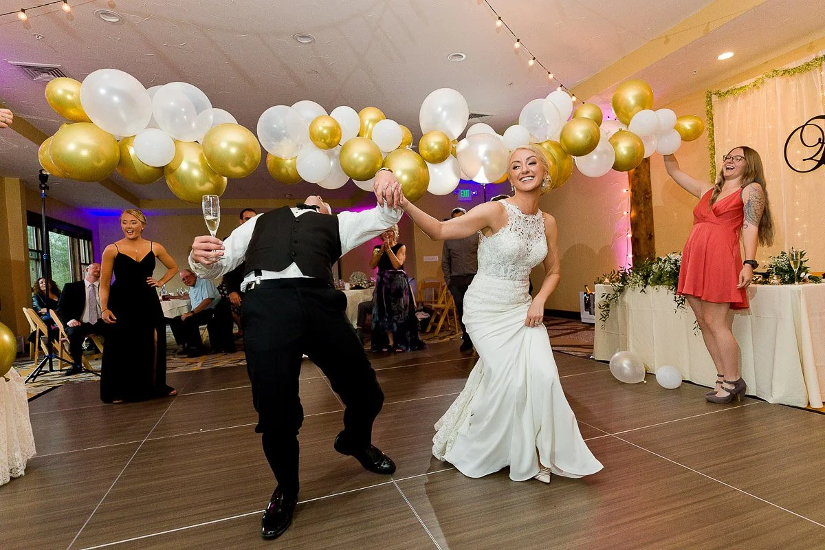 Bride and groom dancing joyfully under gold and white balloons at a wedding reception. Guests smile and celebrate in a festive, elegant setting.