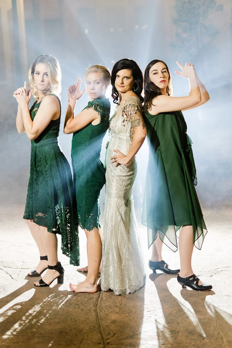 Four women stand back-to-back in style, mimicking a “Charlie's Angels” pose. Three wear green dresses, one silver, with a dramatic, illuminated backdrop.