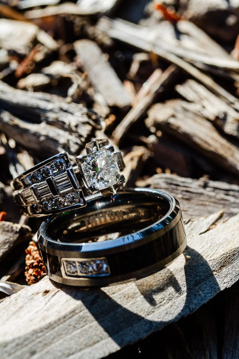 Close-up of two rings on textured wood: a diamond engagement ring and a black band. Sunlight casts shadows, creating a rustic and romantic mood.