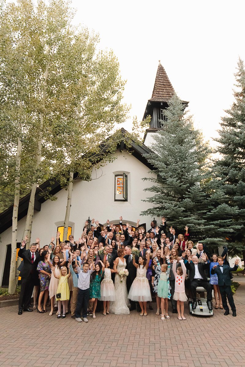 A large, joyful wedding group poses outside a quaint chapel surrounded by trees. People of all ages smile and wave, creating a festive atmosphere.