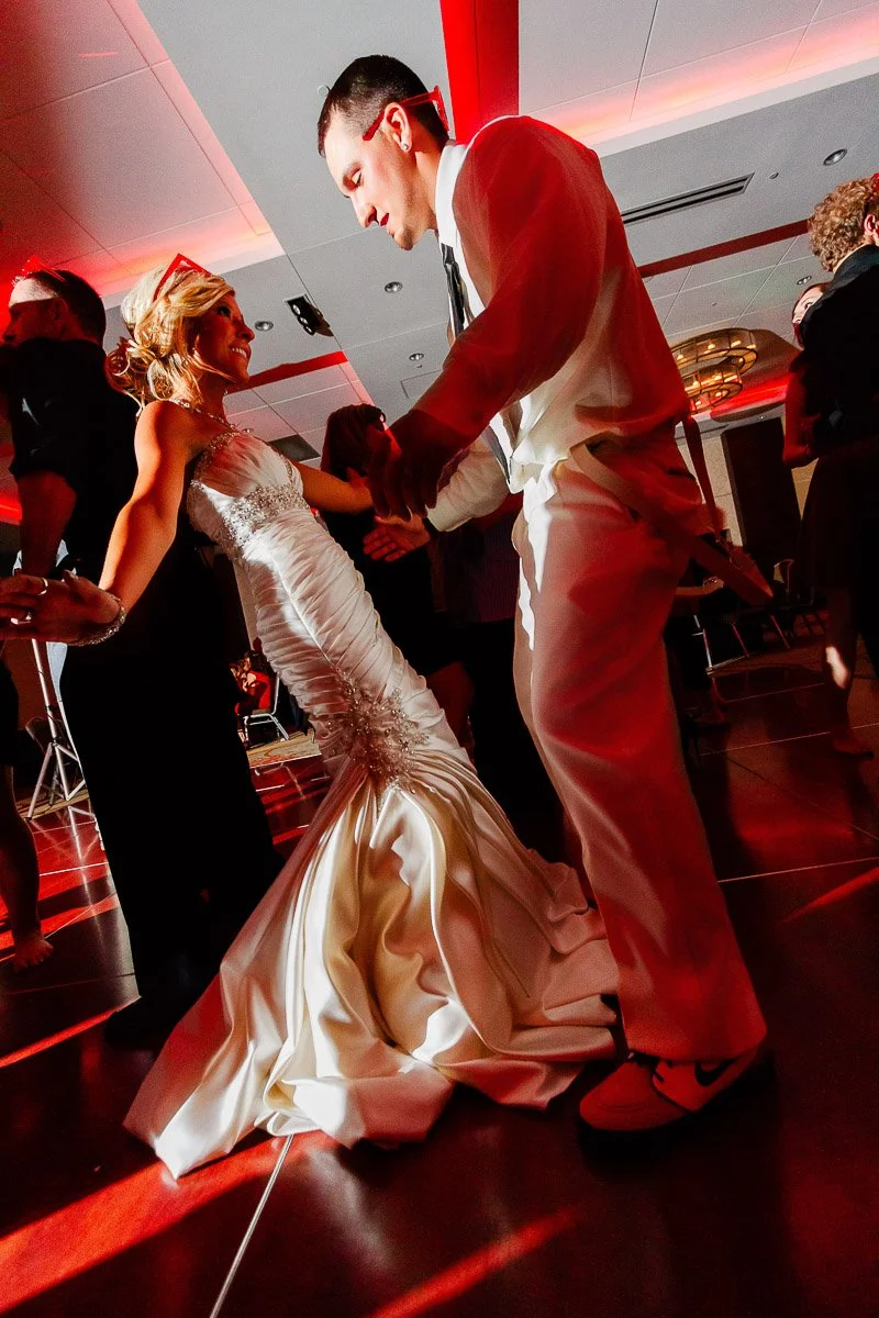 Bride and groom dance joyfully at a wedding, in a warmly lit reception hall. She wears a fitted white gown; he, a white suit with sneakers.