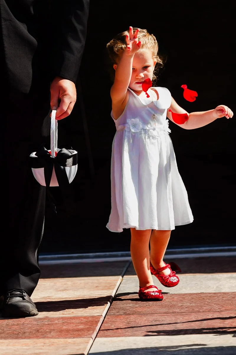 A young girl in a white dress and red shoes enthusiastically tosses flower petals. She holds hands with an adult in a black suit, carrying a basket. Bright and joyful scene.