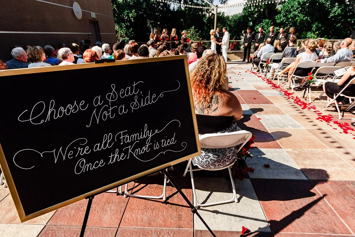 Outdoor wedding ceremony with a chalkboard sign reading "Choose a Seat, Not a Side." Guests sit on either side of rose petal-lined aisle, bride and groom in distance.
