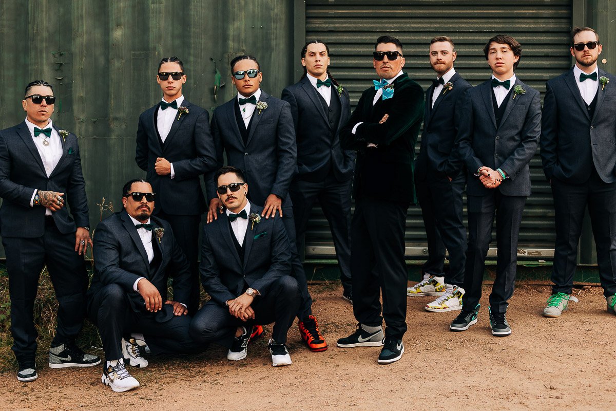 A group of men in formal suits and bow ties pose confidently, wearing colorful sneakers and sunglasses. They stand and crouch in front of a corrugated metal wall, exuding a stylish and playful vibe.