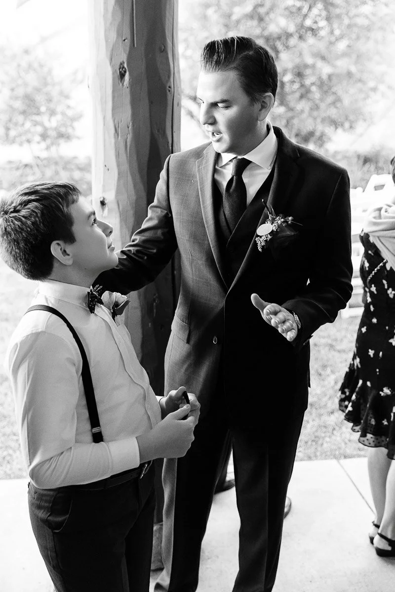 A man in a suit talks to a boy in suspenders and a bow tie. The setting is outdoors. Their expressions are engaged and sincere, conveying a meaningful conversation.