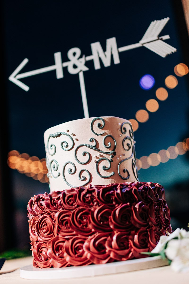 Two-tiered wedding cake with swirled icing patterns. The top tier is white with green swirls; the bottom is deep red roses. A white arrow topper displays "I & M."