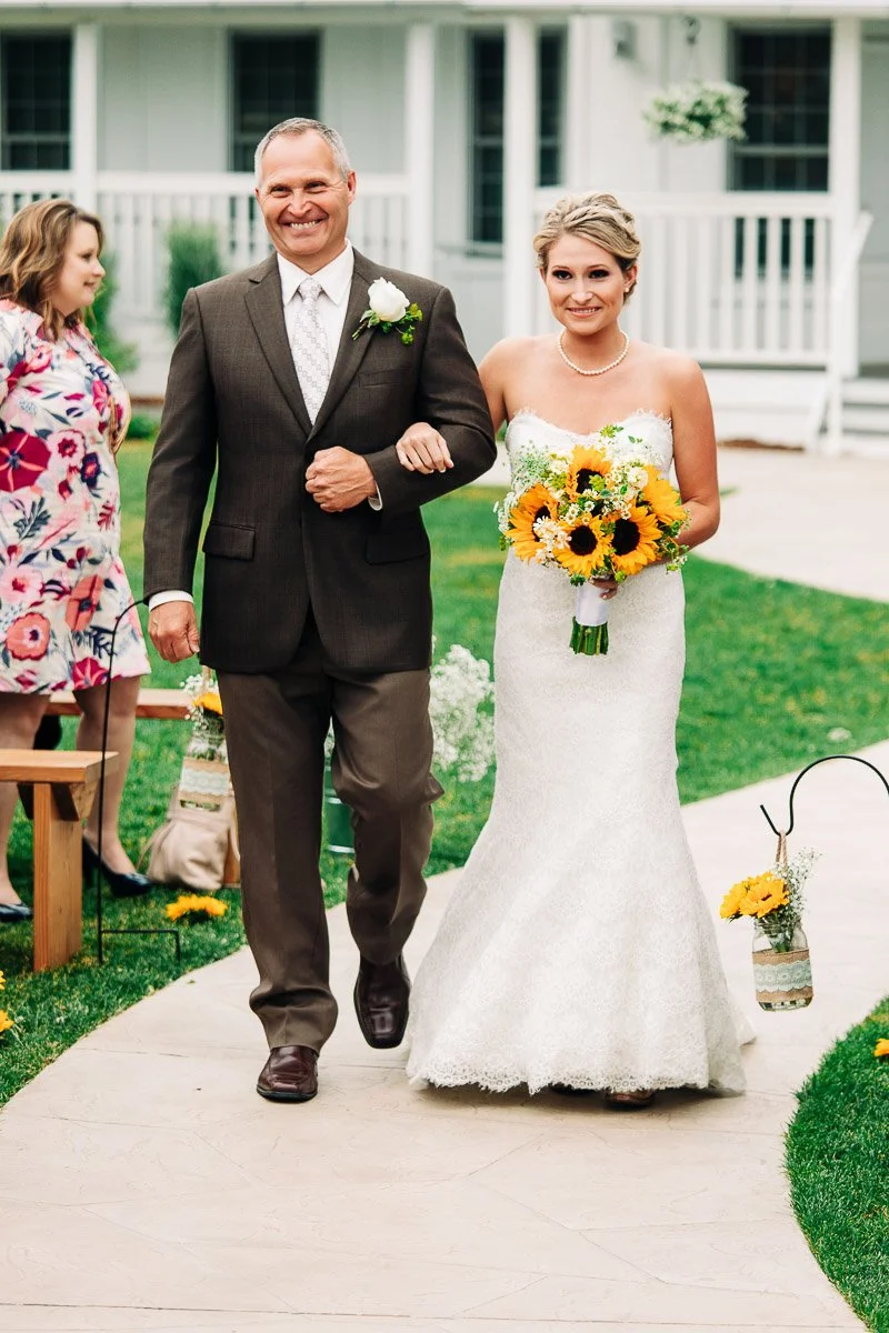 A smiling bride in a strapless lace gown and pearl necklace walks arm-in-arm with a man in a brown suit. She holds a bouquet of sunflowers.