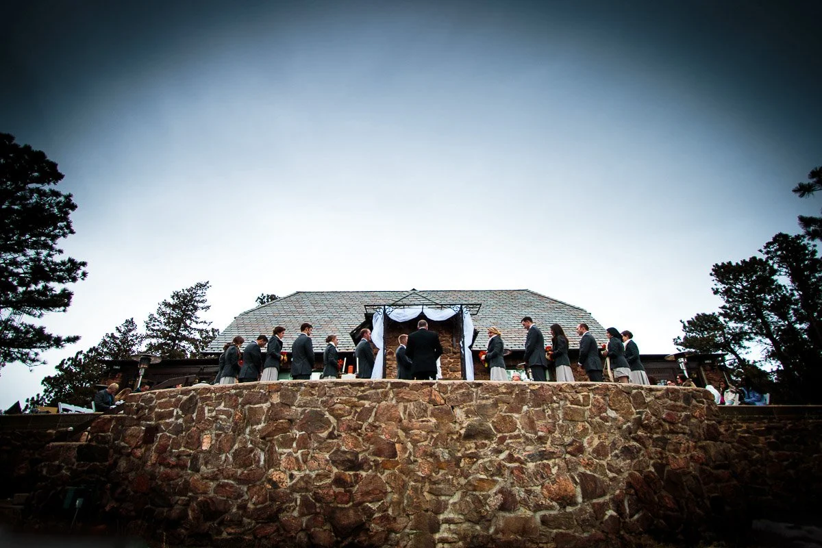 A Boettcher Mansion wedding ceremony on a stone terrace under a cloudy sky. The couple stands at the altar, surrounded by a wedding party in formal attire, conveying a serene and intimate atmosphere.