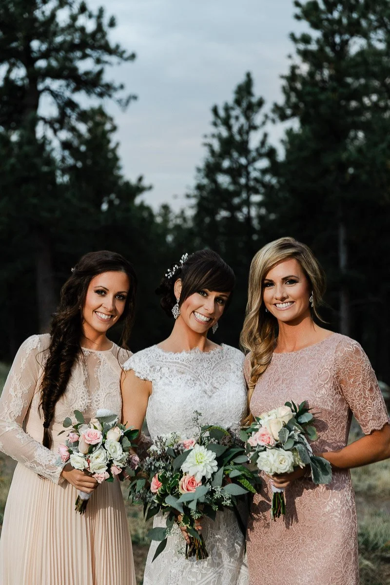 Three smiling women in elegant lace dresses hold bouquets with pink and white flowers. They're outdoors, surrounded by tall pine trees at dusk.