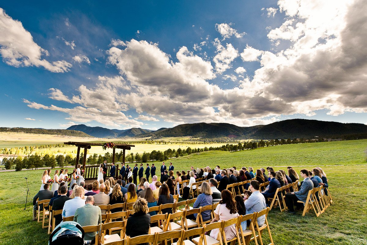 A scenic outdoor Spruce Mountain Ranch wedding ceremony under a vibrant blue sky with fluffy clouds. Guests seated on wooden chairs, surrounded by lush green fields and mountains.