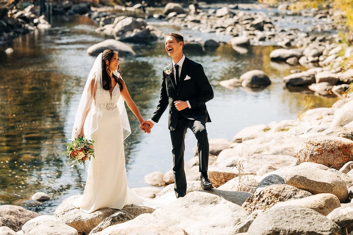 A joyful bride and groom stand on rocky terrain by a serene river, holding hands. She wears a lace gown and he a black suit. Sunlight filters through, creating a warm, romantic scene.