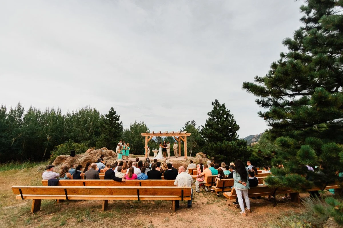 Outdoor wedding ceremony with guests seated on wooden benches facing a wooden arbor. The scene is set in a natural, forested environment.