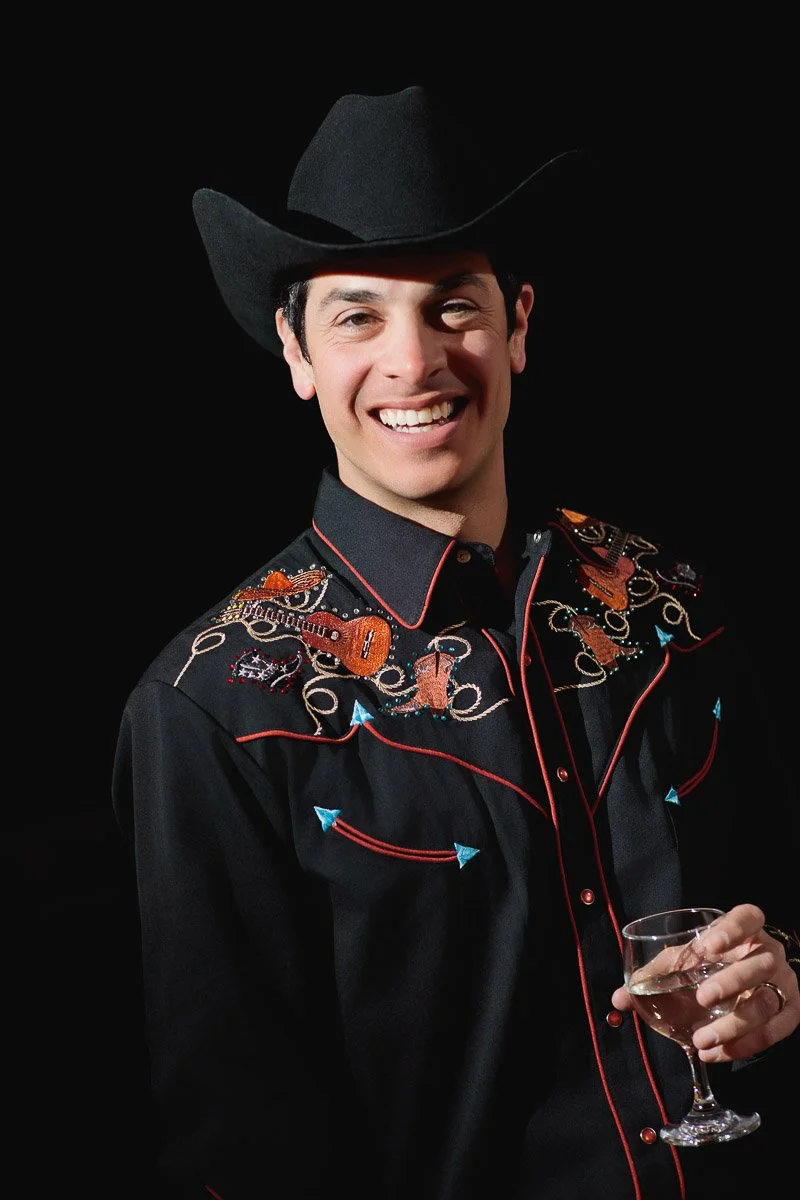 Smiling man in black cowboy hat and embroidered shirt holds a glass of wine, standing against a dark background, exuding a cheerful, festive vibe.