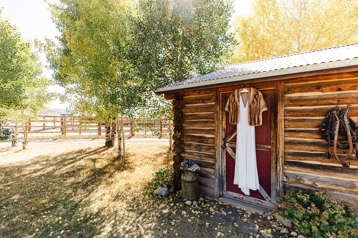 A rustic log cabin with a red door is surrounded by autumn trees. A white dress and fur stole hang on the door, evoking a serene, nostalgic mood.