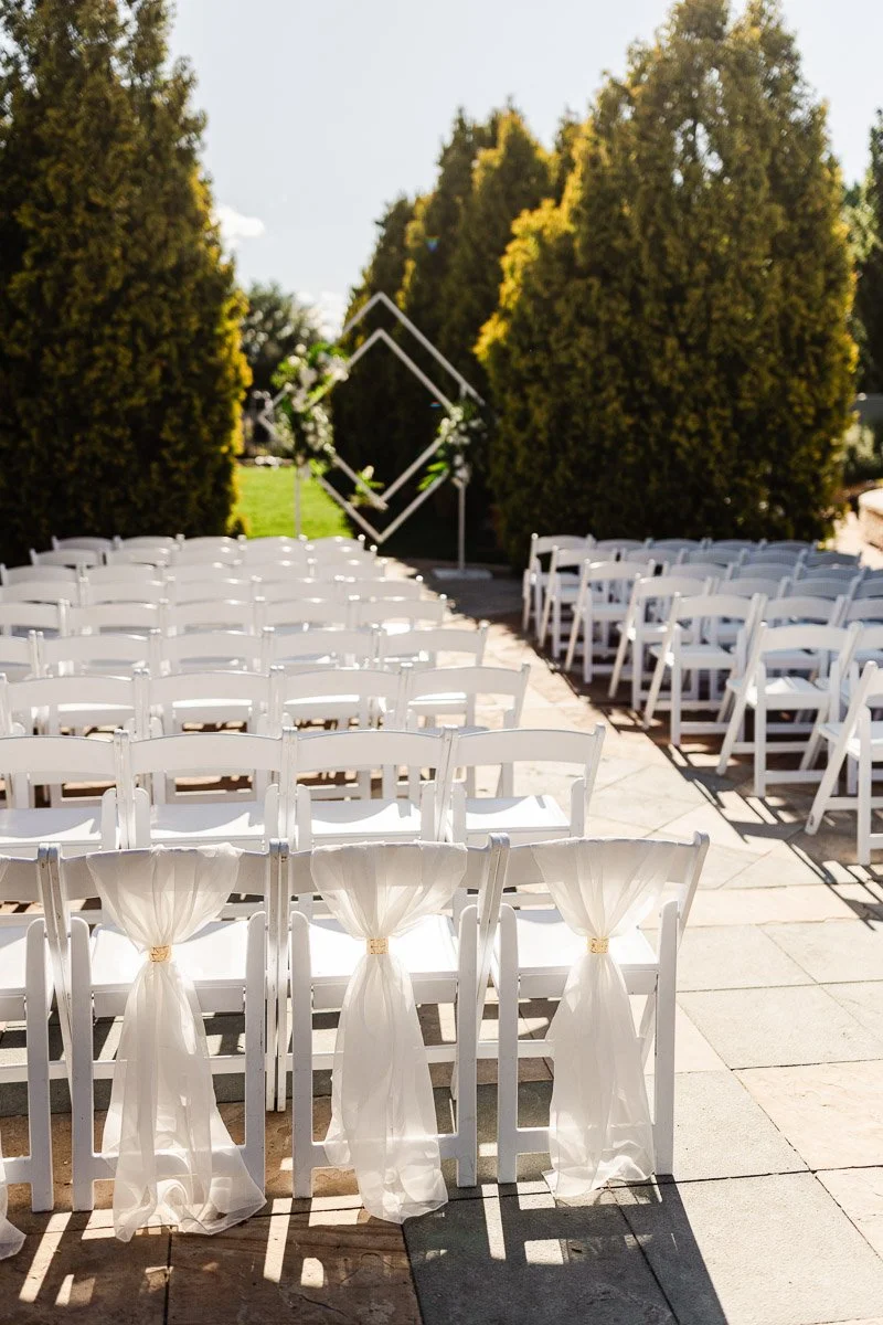 Outdoor wedding setup with rows of white chairs adorned with sheer fabric, facing a geometric arch framed by tall, sunlit trees. Tranquil and elegant before a York Street Botanic Gardens wedding in Denver, Colorado