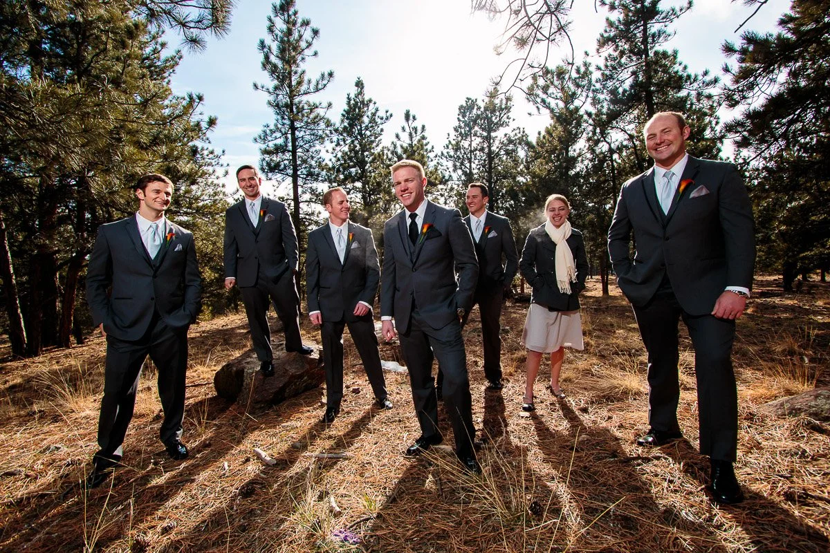 Seven people in formal attire stand together in a sunlit forest at a Boettcher Mansion wedding. The group, mostly smiling, conveys a joyful, celebratory mood amidst tall pine trees.