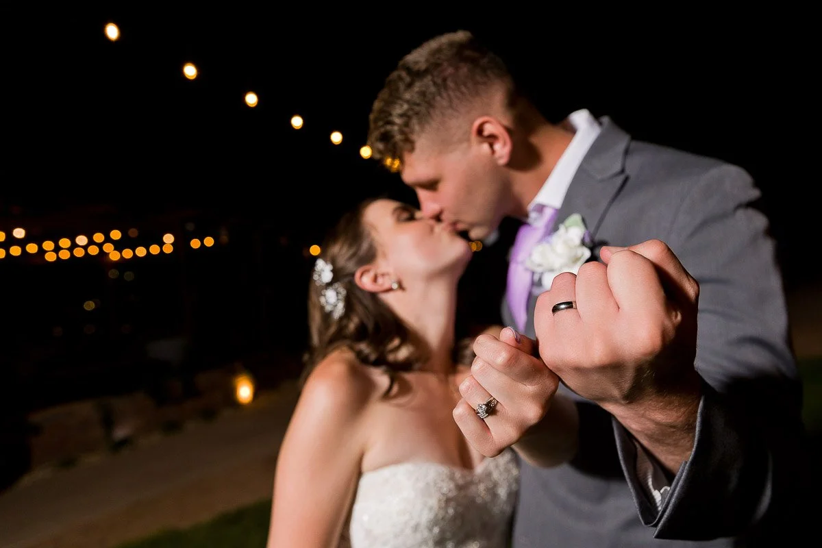 Bride and groom kiss under string lights at night, displaying their wedding rings. The scene is romantic and joyful, emphasizing love and celebration.