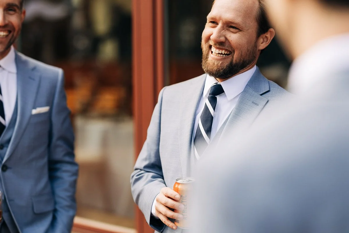 A man in a light blue suit, holding a drink can, smiles warmly while chatting with others in similar attire. The scene conveys a joyful, celebratory mood.